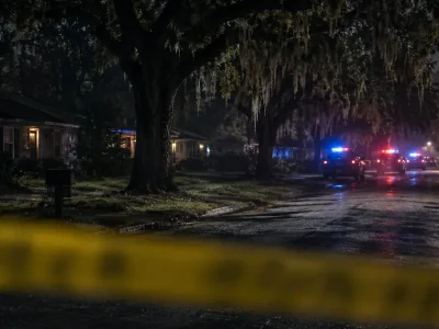 Yellow police barrier tape blurred across the foreground of a dark residential street lined with moss-draped live oaks, with patrol vehicle emergency lights glowing further down the road
