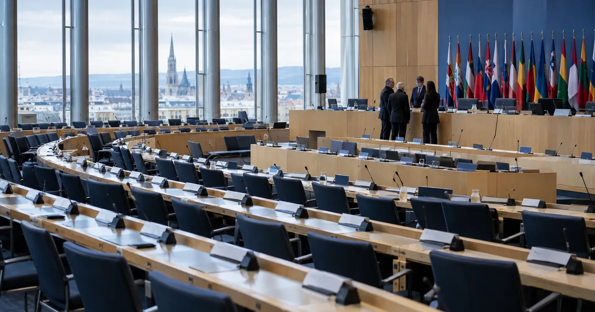 Rows of empty delegate desks in a tiered assembly chamber, with a small group of officials talking beside a line of national flags and a city skyline through tall windows