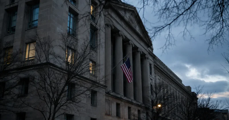 The neoclassical limestone facade of the United States Department of Justice headquarters in Washington at dusk, an American flag hanging between the tall columns and bare winter branches in the foreground.