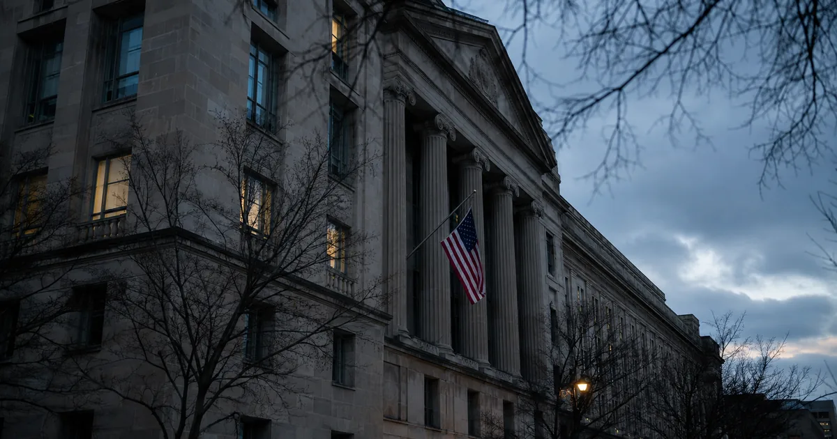 The neoclassical limestone facade of the United States Department of Justice headquarters in Washington at dusk, an American flag hanging between the tall columns and bare winter branches in the foreground.