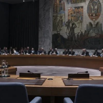The United Nations Security Council chamber seen from behind two empty delegate seats with folded microphones, other members seated around the horseshoe table