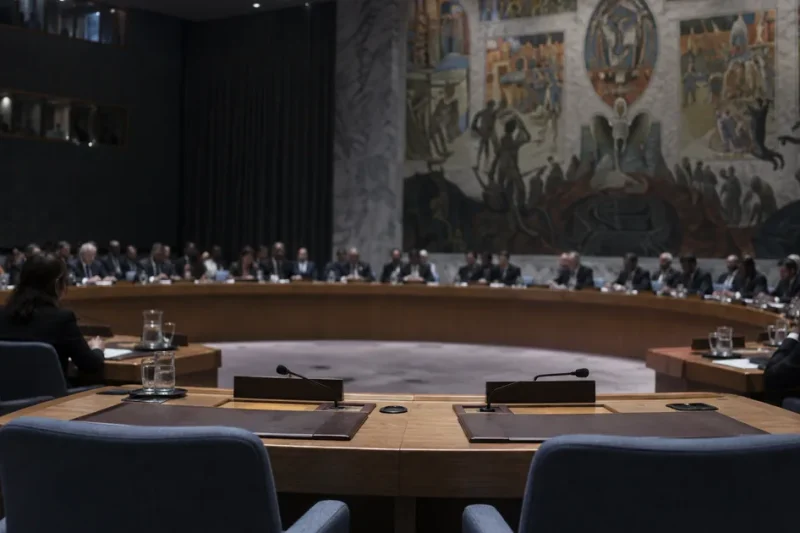 The United Nations Security Council chamber seen from behind two empty delegate seats with folded microphones, other members seated around the horseshoe table