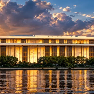 The Kennedy Center's white marble colonnade glowing gold in late sunlight above the Potomac, under a dramatic broken sky