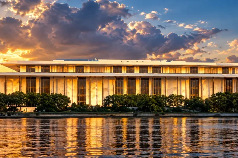 The Kennedy Center's white marble colonnade glowing gold in late sunlight above the Potomac, under a dramatic broken sky