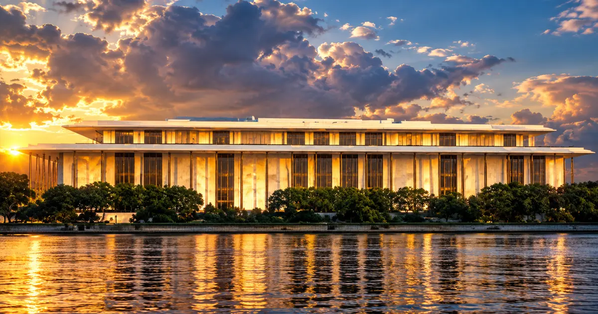 The Kennedy Center's white marble colonnade glowing gold in late sunlight above the Potomac, under a dramatic broken sky