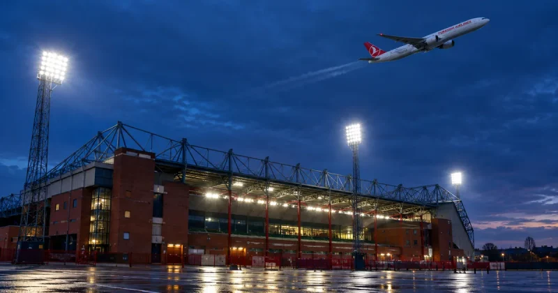A Turkish Airlines passenger jet banks across a deep blue evening sky above the floodlit exterior of a large English football stadium