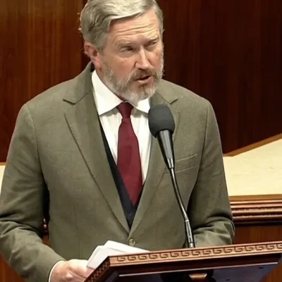 Representative Thomas Massie, grey-bearded in an olive tweed jacket and dark red tie, speaks into a microphone at the wooden House of Representatives lectern while holding papers