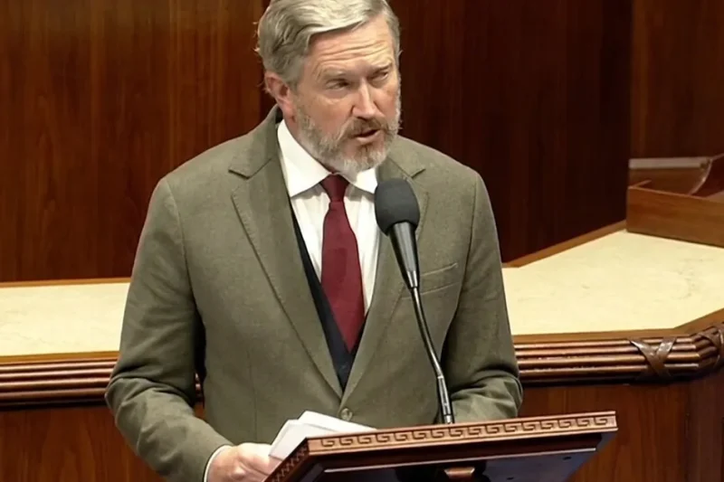 Representative Thomas Massie, grey-bearded in an olive tweed jacket and dark red tie, speaks into a microphone at the wooden House of Representatives lectern while holding papers