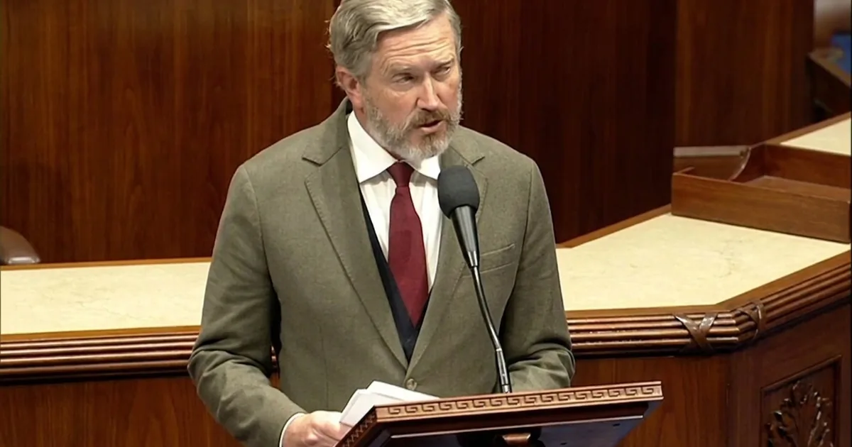 Representative Thomas Massie, grey-bearded in an olive tweed jacket and dark red tie, speaks into a microphone at the wooden House of Representatives lectern while holding papers