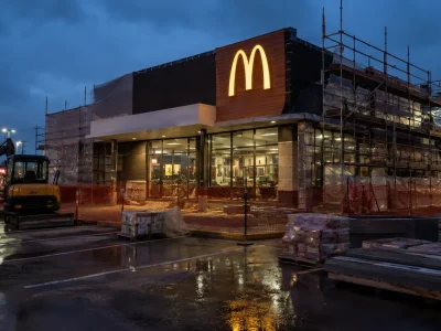 A McDonald's restaurant wrapped in scaffolding and orange safety fencing during a renovation, lit golden arches above a wet parking lot at dusk