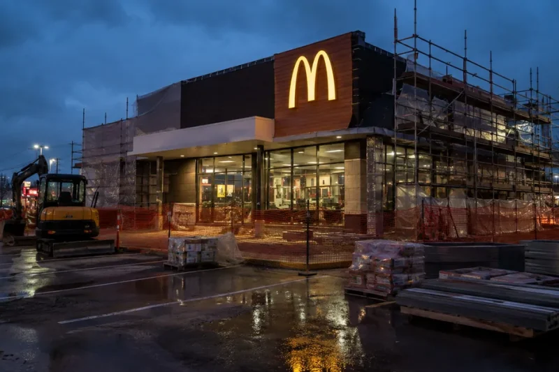 A McDonald's restaurant wrapped in scaffolding and orange safety fencing during a renovation, lit golden arches above a wet parking lot at dusk