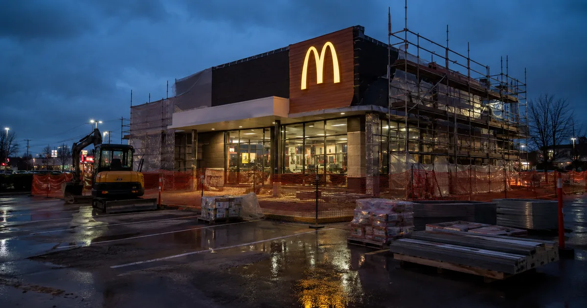 A McDonald's restaurant wrapped in scaffolding and orange safety fencing during a renovation, lit golden arches above a wet parking lot at dusk