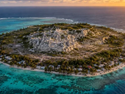 Aerial photograph of a small coral island at golden hour, a thin coastal ring of houses and palm trees surrounding a raised interior plateau of bare white coral pinnacles, ringed by turquoise reef and open ocean