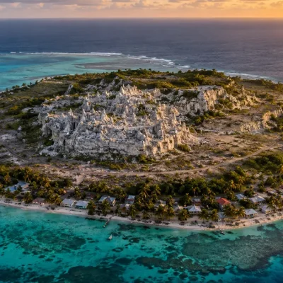 Aerial photograph of a small coral island at golden hour, a thin coastal ring of houses and palm trees surrounding a raised interior plateau of bare white coral pinnacles, ringed by turquoise reef and open ocean
