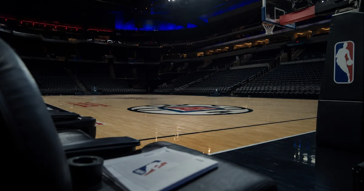 A darkened empty basketball arena, the Los Angeles Clippers logo at center court and an NBA-branded stanchion pad courtside