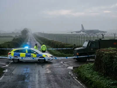 Police car and army bomb disposal truck behind cordon tape on a misty country road beside an airfield fence, with a B-52 bomber on the runway beyond