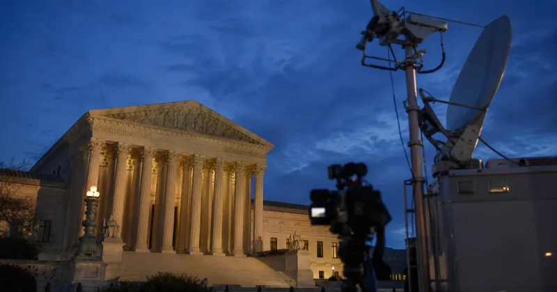 The United States Supreme Court building lit by floodlights at dusk, with the dish and mast of a television news satellite truck and a broadcast camera silhouetted in the foreground.