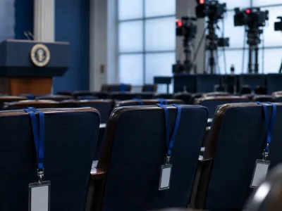 Three empty chairs with press credentials on blue lanyards draped over the seatbacks in the White House briefing room, television cameras on risers behind them