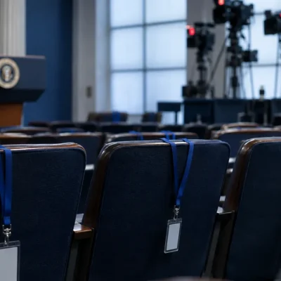 Three empty chairs with press credentials on blue lanyards draped over the seatbacks in the White House briefing room, television cameras on risers behind them
