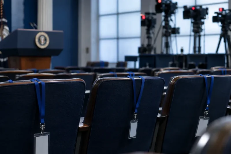 Three empty chairs with press credentials on blue lanyards draped over the seatbacks in the White House briefing room, television cameras on risers behind them