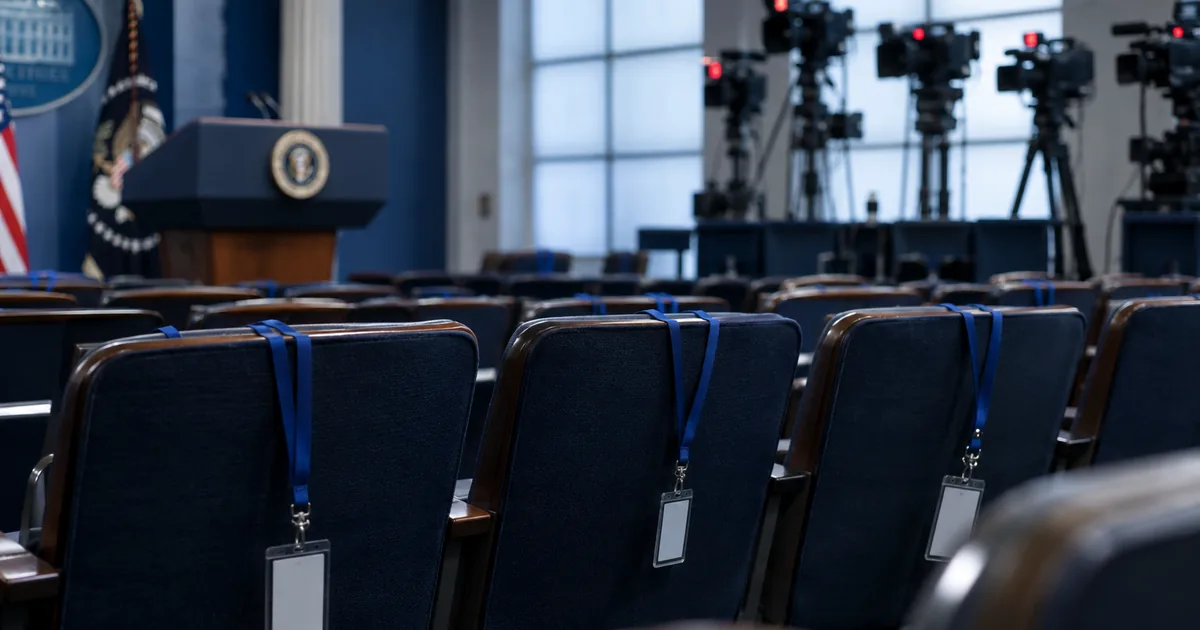 Three empty chairs with press credentials on blue lanyards draped over the seatbacks in the White House briefing room, television cameras on risers behind them