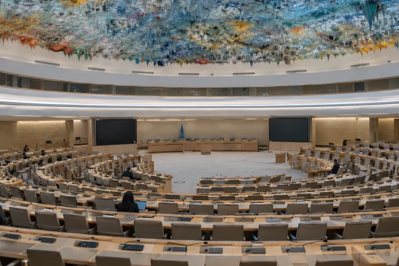Rows of empty curved delegate desks beneath the colorful stalactite ceiling of the UN Human Rights Council chamber in Geneva