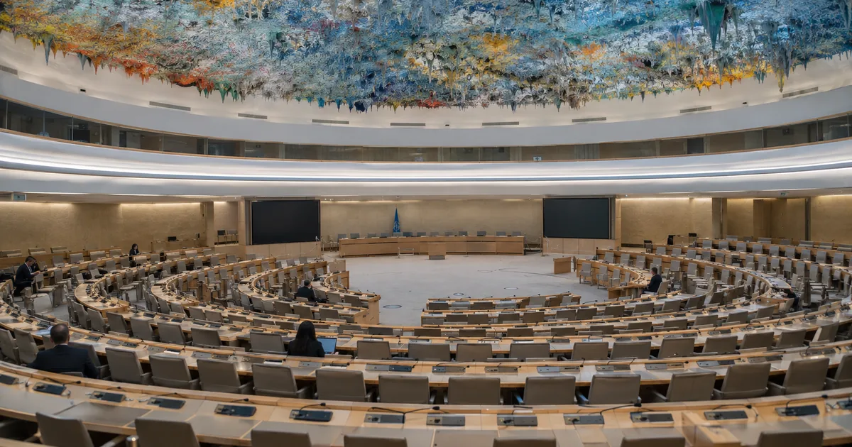 Rows of empty curved delegate desks beneath the colorful stalactite ceiling of the UN Human Rights Council chamber in Geneva