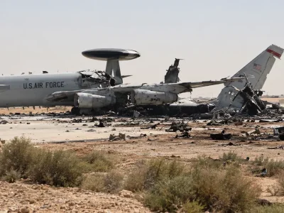 Illustration of a gray U.S. Air Force E-3 Sentry radar plane on a desert air base apron with its rear fuselage and tail torn apart, debris and a burned truck nearby