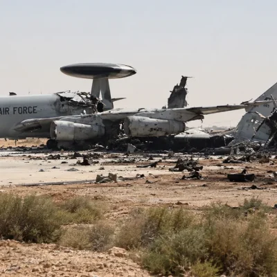 Illustration of a gray U.S. Air Force E-3 Sentry radar plane on a desert air base apron with its rear fuselage and tail torn apart, debris and a burned truck nearby
