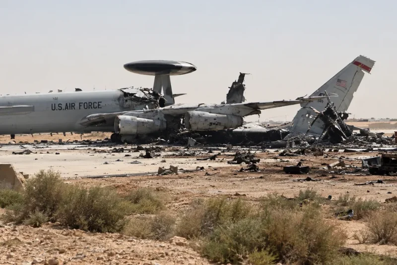 Illustration of a gray U.S. Air Force E-3 Sentry radar plane on a desert air base apron with its rear fuselage and tail torn apart, debris and a burned truck nearby