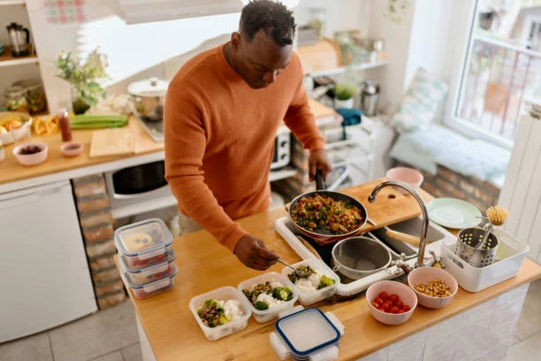 A man portions a vegetable and grain skillet into stacked meal prep containers on a kitchen island