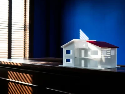 A white architectural model of a house with a red roof sits on a dark wooden counter in front of a cobalt blue wall, with striped shadows from window blinds across the wood.