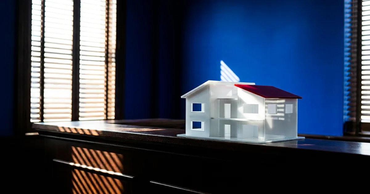 A white architectural model of a house with a red roof sits on a dark wooden counter in front of a cobalt blue wall, with striped shadows from window blinds across the wood.