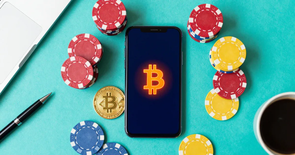 Overhead view of a phone showing a glowing Bitcoin symbol surrounded by stacks of poker chips and a gold Bitcoin coin on a teal desk