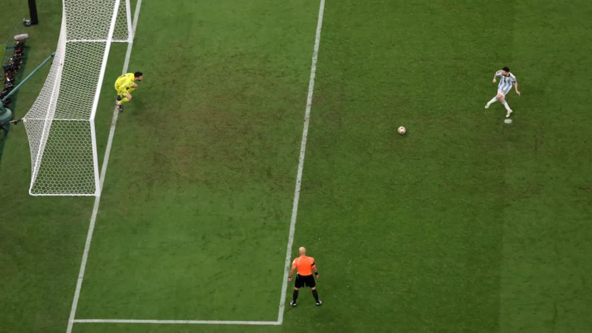 Aerial view of a penalty kick with the goalkeeper diving early and the referee watching from the edge of the box