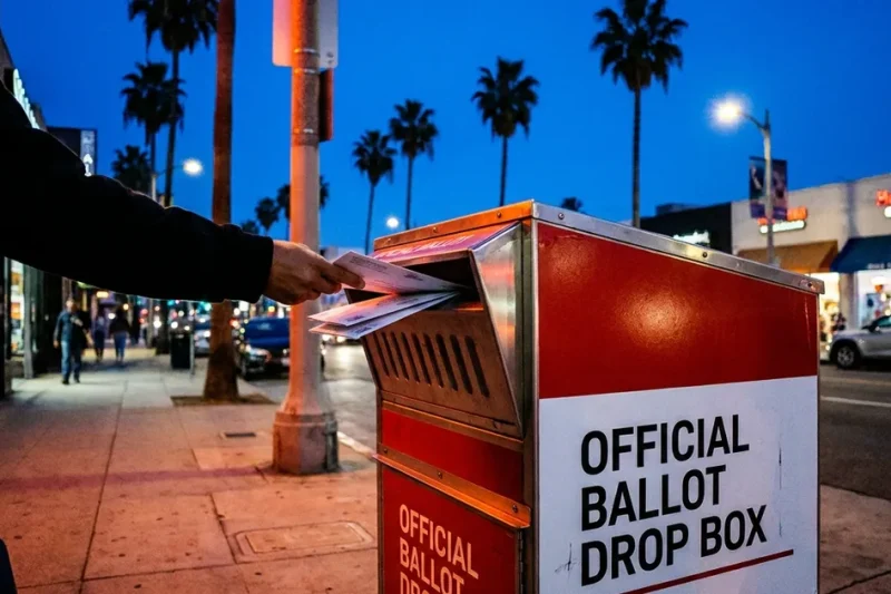 A hand pushes mail ballot envelopes into a red and white official ballot drop box on a Los Angeles sidewalk at dusk, palm trees behind it