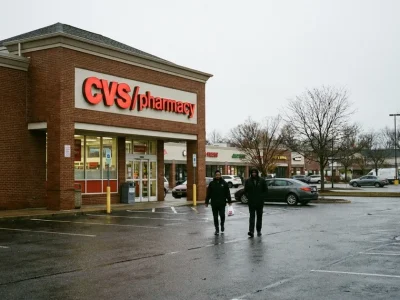 Two shoppers walk across a wet parking lot from a CVS Pharmacy store on an overcast autumn day