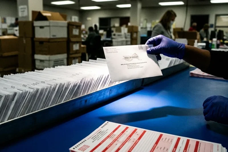 A gloved election worker lifts one mail ballot envelope out of a long tray of envelopes at a blue table