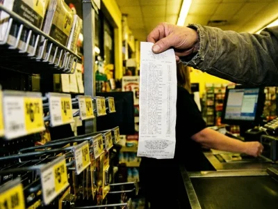 A shopper holds up a long paper receipt at a checkout counter beside shelves of yellow price tags