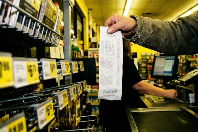 A shopper holds up a long paper receipt at a checkout counter beside shelves of yellow price tags