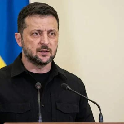 Volodymyr Zelensky in a black field shirt speaks at a lectern with two microphones, a Ukrainian flag behind him
