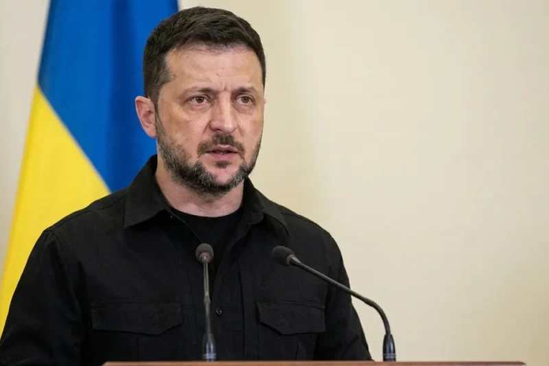 Volodymyr Zelensky in a black field shirt speaks at a lectern with two microphones, a Ukrainian flag behind him