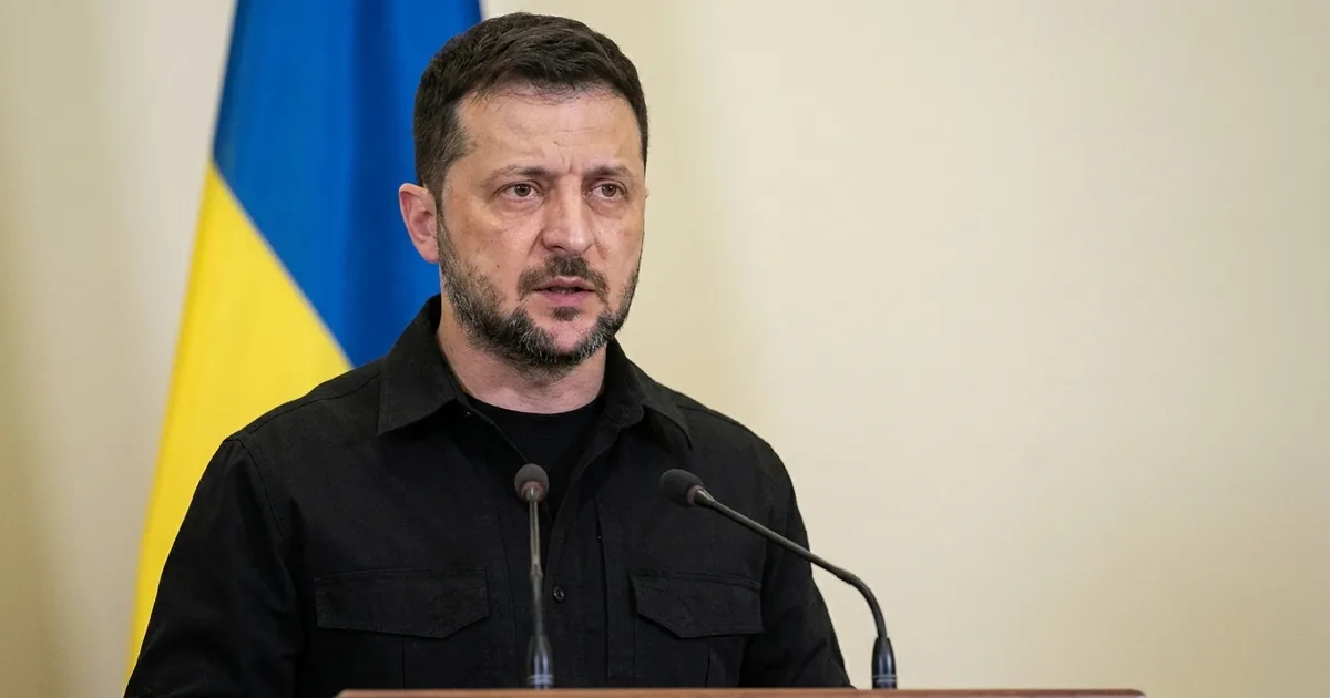 Volodymyr Zelensky in a black field shirt speaks at a lectern with two microphones, a Ukrainian flag behind him