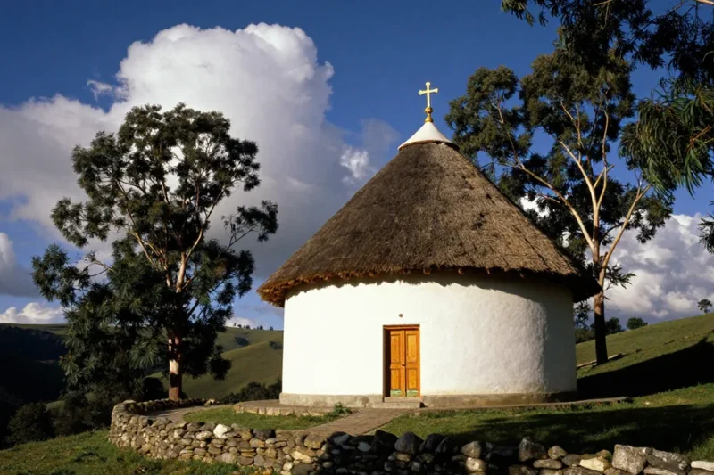 Illustrative image of a round whitewashed Ethiopian Orthodox church with a thatched roof and a gold cross on a green highland hillside under a deep blue sky