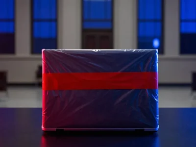 A closed laptop sealed in a clear evidence bag with red tape on a dark table in a courtroom lit blue and red