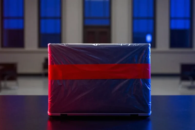 A closed laptop sealed in a clear evidence bag with red tape on a dark table in a courtroom lit blue and red