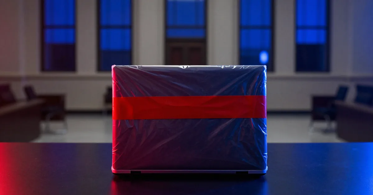 A closed laptop sealed in a clear evidence bag with red tape on a dark table in a courtroom lit blue and red