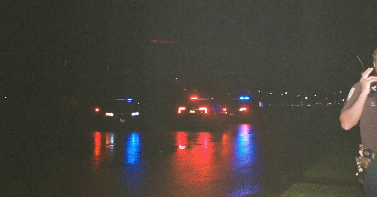 Police cars with flashing red and blue lights reflected on a wet street at night, an officer speaking into a shoulder radio at the right edge of the frame