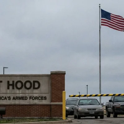 The Fort Hood entrance sign and guard booth under an overcast sky, with a military police soldier at the gate and cars waiting at the barrier