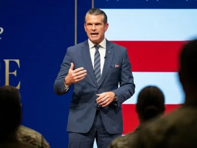 Defense Secretary Pete Hegseth in a navy suit speaks on stage in front of an American flag backdrop, with service members in the foreground